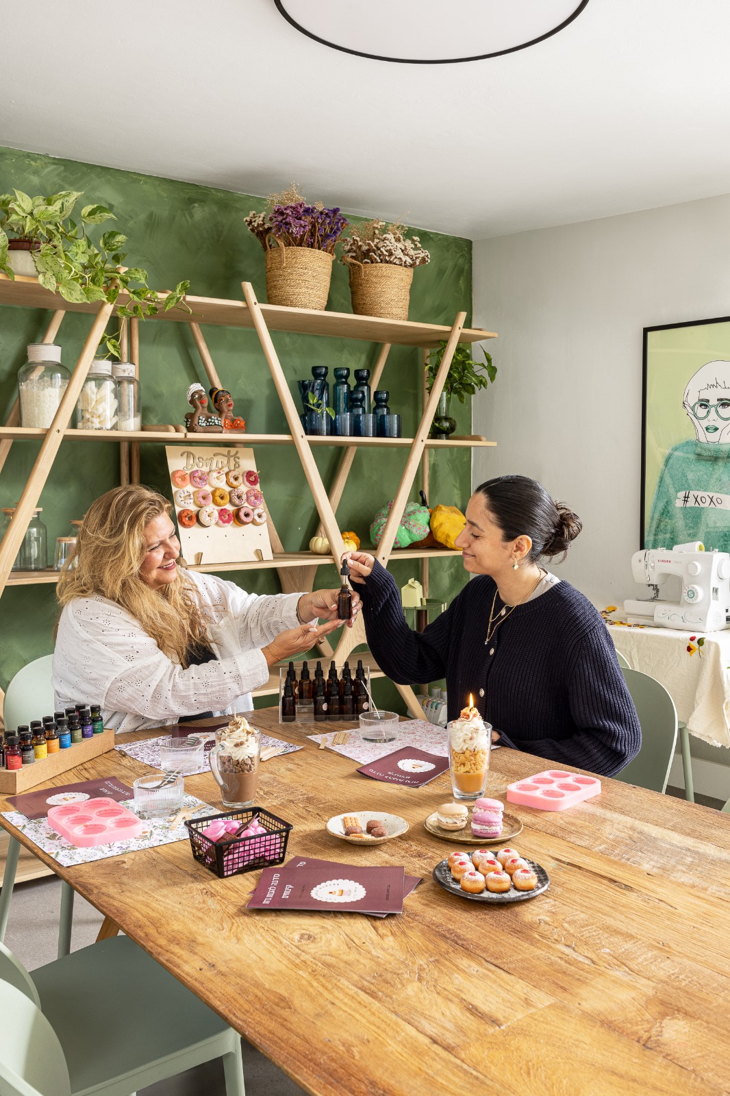 שתי נשים יוצרות ביחד בסטודיו של ArtyParty בתל מונד — שולחן עץ, מדפי יצירה, קיר ירוק